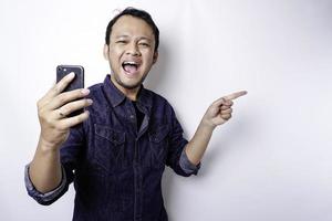 homem asiático animado vestindo camisa azul apontando para o espaço da cópia ao lado dele enquanto segura o telefone, isolado pelo fundo branco foto