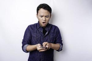 homem asiático chocado vestindo camisa azul e segurando seu telefone, isolado por fundo branco foto