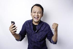 um jovem asiático com uma expressão feliz e bem-sucedida vestindo camisa azul e segurando seu telefone, isolado por fundo branco foto