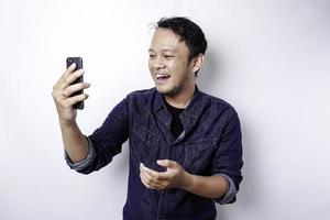 homem asiático animado vestindo camisa azul apontando para o espaço da cópia ao lado dele enquanto segura o telefone, isolado pelo fundo branco foto