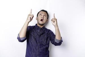 homem asiático chocado vestindo camisa azul apontando para o espaço da cópia em cima dele, isolado pelo fundo branco foto