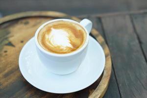 café quente com espuma floral em um copo branco fundo de madeira cinza foto