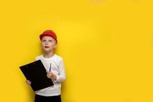 menino em um capacete de segurança de construção com um tablet e uma caneta na mão olha com espanto em um fundo amarelo com espaço de cópia foto