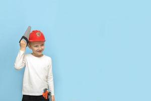 menino com uma serra de brinquedo experimenta um capacete de segurança de construção em um fundo azul com espaço de cópia foto
