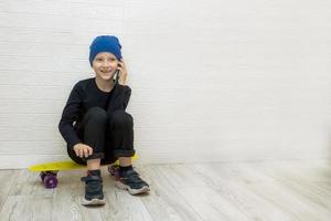 menino de chapéu sentado em um skate conversando alegremente ao telefone contra uma parede branca com espaço de cópia foto