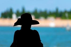 silhueta de turista com chapéu de sol olha para a lagoa veneziana em fundo desfocado foto
