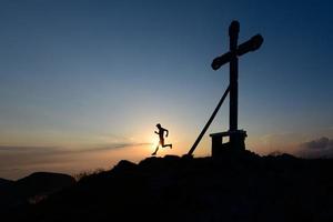 silhueta de homem correndo sob o sol poente no topo de uma montanha com a cruz foto
