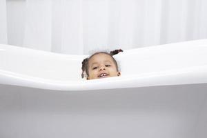 uma menina africana engraçada olha para fora de uma banheira branca e sorri.