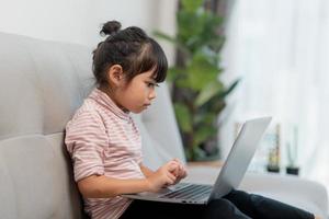 garotinha asiática da escola usa computador portátil sentado no sofá sozinho em casa. criança aprendendo lendo conteúdo de mídia social on-line, jogando lições de educação conversando com amigos. foto