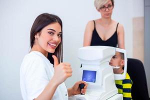 oculista feminina usando máquina para verificar a visão do olho na clínica. garotinho olhando para equipamento e médico testando a pupila ocular na loja ótica. conceito de cuidados com os olhos e saúde. foto