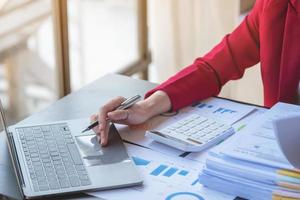 financeira, empresária asiática de terno vermelho segurando a xícara de café sentada na mesa no escritório, tendo computador para fazer trabalho de contabilidade no local de trabalho para calcular o lucro anual por dever, ideia de negócio foto