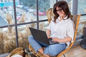 linda empresária trabalhando em um laptop em uma cadeira perto da janela. freelancer feminino conectando-se à internet via computador. blogueiro ou jornalista escrevendo novo artigo. foto