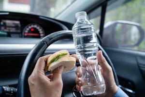 senhora asiática segurando garrafa de água e comida de padaria de pão no carro perigoso e corre o risco de um acidente. foto