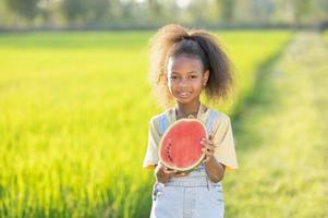 menina bonitinha de pele negra comendo melancia ao ar livre cenário de campo de arroz verde criança africana comendo melancia foto