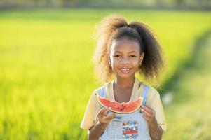 menina bonitinha de pele negra comendo melancia ao ar livre cenário de campo de arroz verde criança africana comendo melancia foto