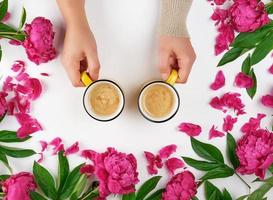 duas mãos segurando xícaras amarelas com uma bebida de café quente em um fundo branco no meio de botões de peônias florescendo foto