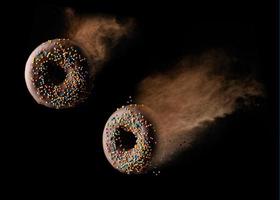 rosquinhas redondas de chocolate com granulado de açúcar multicolorido levitam em uma nuvem de cacau marrom em um fundo preto foto