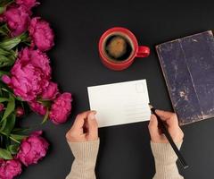 cartão postal branco vazio e duas mãos femininas, sobre a mesa um buquê de peônias vermelhas e uma xícara de café de cerâmica foto