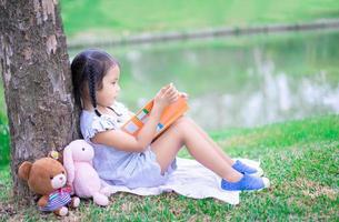 menina bonitinha lendo um livro com uma boneca no parque foto