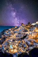 vila de oia na luz do sol com estrelas e via láctea, santorini, grécia. incrível paisagem de férias de verão, arquitetura branca e luzes noturnas. destino de viagem famoso, fundo de viagens urbanas foto