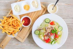 Batatas fritas e ketchup em um prato de madeira e salada com legumes frescos e atum, vista superior com espaço livre para o seu texto. foto