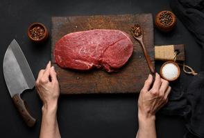 lombo de carne crua está sobre uma tábua de corte de madeira e especiarias para cozinhar em uma mesa preta, vista superior e faca de cozinha grande foto