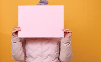 mulher de casaco de inverno rosa e chapéu segura uma folha de papel rosa em branco sobre fundo amarelo. venda sazonal foto