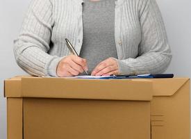 mulher com roupas cinza fica perto de uma caixa marrom e assina documentos com uma caneta. o conceito de movimentação, envio e recebimento de mercadorias foto