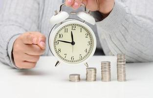 pilha de moedas e despertador branco redondo em cima da mesa. o conceito de tempo é dinheiro, um período de descontos. foto