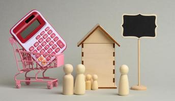 família de figuras de madeira e um corretor de imóveis no fundo de uma casa e uma calculadora em um carrinho em um fundo cinza foto