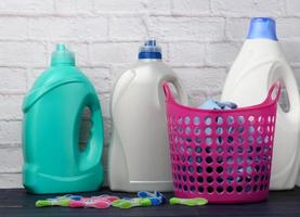 cesto de roupa suja rosa e garrafas de plástico com detergente líquido no fundo da parede de tijolo branco foto