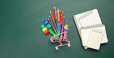 carrinho de compras em miniatura cheio de material de escritório escolar em um quadro de giz verde. preparando-se para a escola, comprando canetas e cadernos foto