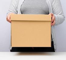 mulher com roupas cinza fica de pé e segura uma caixa marrom em um fundo branco, movendo, enviando e entregando mercadorias foto