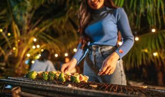 feche a mão da mulher cozinhar carne na churrasqueira na festa de ano novo. bar-bq ou churrasco no fogão tradicional. festa noturna, pessoas e conceito de celebração. foto