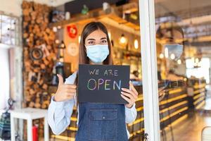 garçonete feliz com máscara facial protetora segurando sinal aberto enquanto está na porta do café ou restaurante, abra novamente após o bloqueio devido ao surto de coronavírus covid-19. mostrando sinal de ok foto