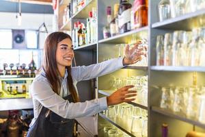 barman colocando vidro limpo na prateleira. jovem mulher atrás do bar tomando um copo limpo para servir uma bebida. barman no bar ou restaurante embalando copos