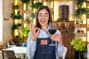 garçonete elegante alegre segurando copo de vinho tinto mostrando sinal delicioso, de pé no restaurante. vinho de degustação de sommelier no restaurante. foto