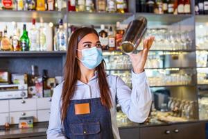 jovem trabalhadora na mesa do barman no restaurante bar preparando coquetel com agitador. mulher jovem e bonita atrás do bar usando máscara facial protetora foto