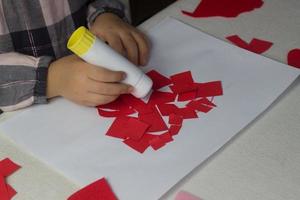 menina fazendo apliques de coração vermelho com papel e cola na folha branca, cartão de dia dos namorados, presente de feriado para o dia das mães, presentes feitos à mão para crianças, símbolo de amor com papel, atividade infantil foto