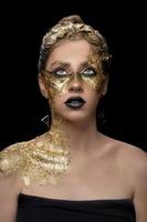 retrato do close up da mulher jovem e bonita com folha de ouro no rosto. maquiagem dourada criativa em fundo preto foto