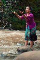 uma linda mulher asiática lavando a mão e brincando com a água em pé perto do rio em um vestido roxo tradicional e saia verde foto