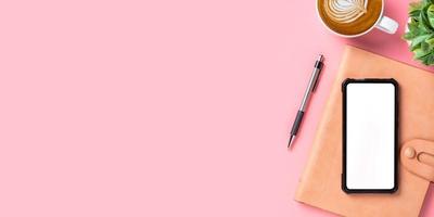 mesa de escritório com telefone inteligente de tela em branco, caneta, caderno, xícara de café no fundo rosa, vista superior com espaço de cópia, mock up. foto