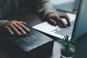 conceito de tecnologia online cristã. mãos orando de cristão com laptop de computador digital, igreja ao vivo online para o culto de domingo. a mulher católica asiática está lendo o livro da bíblia sagrada e o estudo online. foto