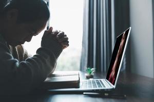 conceito de tecnologia online cristã. mãos orando de cristão com laptop de computador digital, igreja ao vivo online para o culto de domingo. a mulher católica asiática está lendo o livro da bíblia sagrada e o estudo online. foto