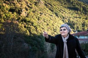 mulher idosa de jaqueta preta e chapéu apontando para as montanhas foto