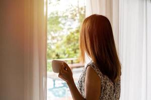 jovem mulher com uma xícara de café abrindo as cortinas pela manhã e olhando pela janela foto
