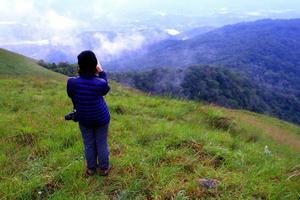 turista ou caminhadas no suéter azul com chapéu preto e segurando a câmera sozinho no campo de grama verde com ar fresco e névoa no fundo da montanha com espaço de cópia, vista de paisagem de beleza e viagens foto