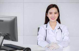médica profissional asiática sorridente sentada na sala de exames no hospital enquanto está na mesa com papel e computador no conceito de saúde foto