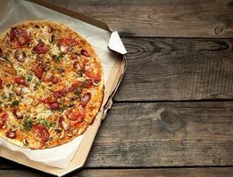 pizza redonda assada com linguiça defumada, cogumelos, tomate, queijo e endro em uma caixa de papelão aberta sobre uma mesa de madeira foto