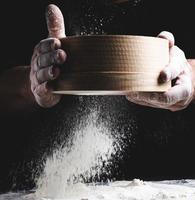 peneira de madeira redonda com farinha nas mãos masculinas, o chef peneira a farinha de trigo branca foto